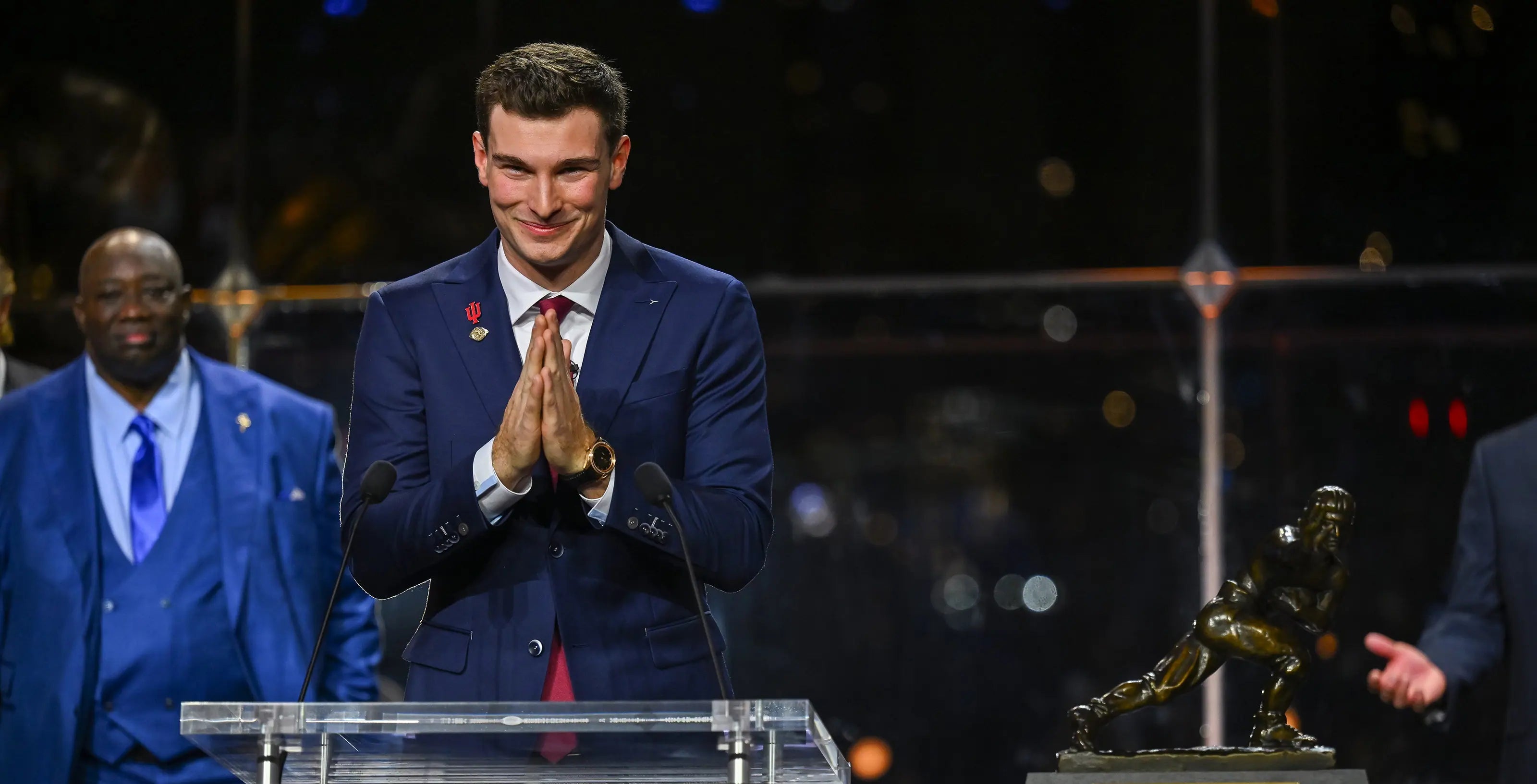Man in a suit standing behind a Heisman Trophy podium with the trophy in the background.